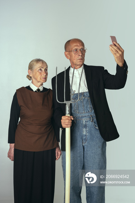 Elderly man and woman in art performance, replica of painting american gothic. Retro style, comparison of eras and cultural, humor concept.
