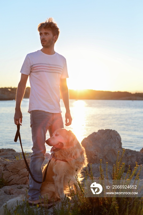 Handsome adult is enjoying the sunset with his dog together by the ocean. Golden Retriever and his human have a great friendship.