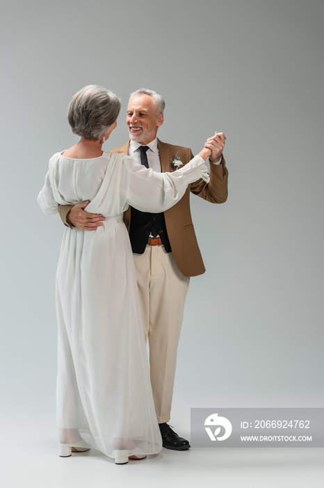 full length of happy middle aged groom and bride in wedding dress holding hands while dancing on grey