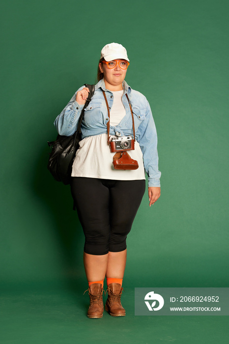 Portrait of emotive young woman in casual clothes, cap, with camera and backpack posing over green studio background. Travel time, vacation