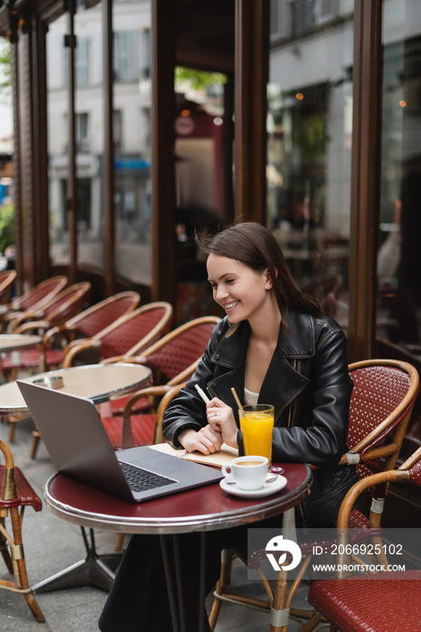 happy freelancer in black leather jacket looking at laptop near drinks on table in french outdoor cafe.