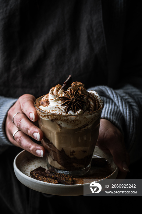 Woman in sweater and dark apron holding plate with  glass of Pumpkin pie spice mocha latte with whipped cream  and dark chocolate  on black background .