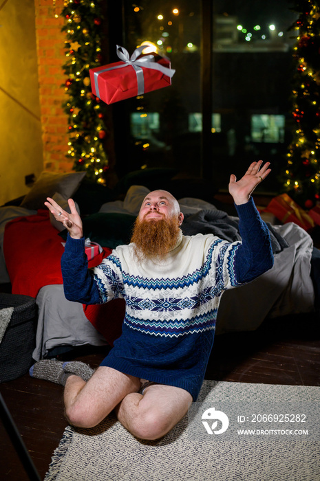 A man with a long red beard sits on the floor without pants in a winter sweater. man is holding a wrapped present on the background of New Year decorations and lights. Christmas tree. Parody, humor.