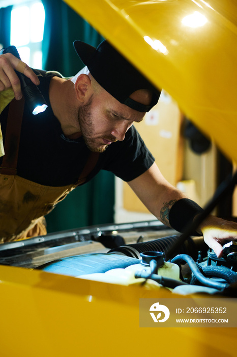 Serious auto mechanic with flashlight bending over open hood of yellow car, checking defects and fixing parts of motor in garage