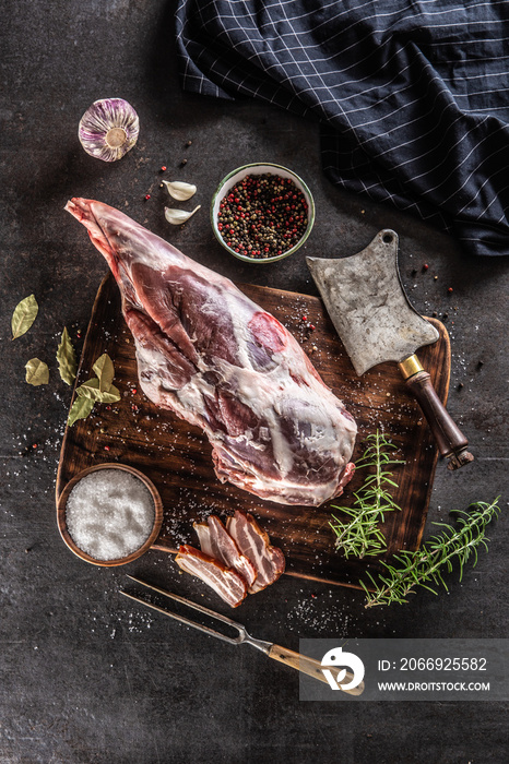 Raw lamb thigh surrounded by salt, pepper, garlic, bacon and rosemary on a vinateg surface