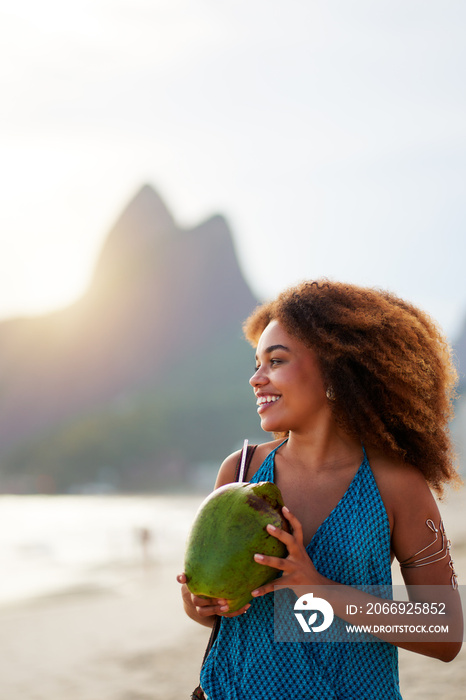 brazilian black woman holds a coconut in her hands on the beach with mountain two brothers in the background at sunset in Ipanema