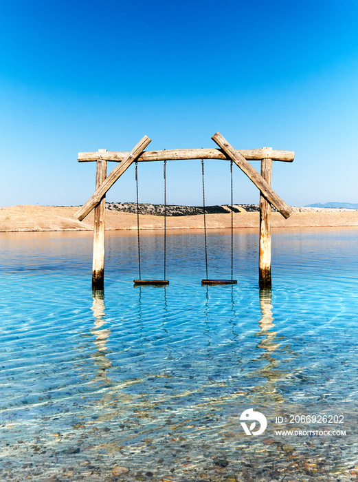 Empty swing in the water. Pebble beach on Adriatic coast of Croatia. Island Krk on background. Jadranovo village near Rijeka in Dalmatia, Croatia. Romantic wooden swing in transparent sea water.