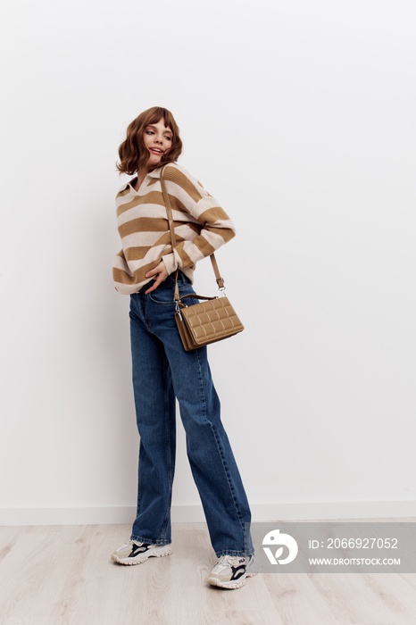 a full-length photo of stylish woman standing on a light background in a striped sweater and blue jeans, posing relaxed with a beige bag worn over her shoulder, adjusting the sweater and looking away