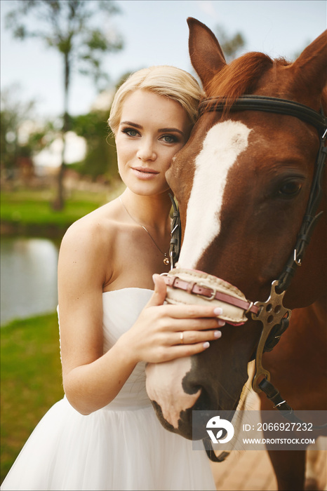 Beautiful and fashionable young bride, blonde model girl with blue eyes and stylish hairstyle in white dress posing with brown horse outdoors