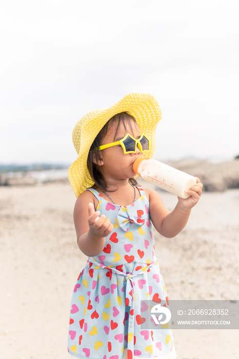 Portrait of Asian baby girl wearing hat and sunglasses sucking milk from bottle on the beach.