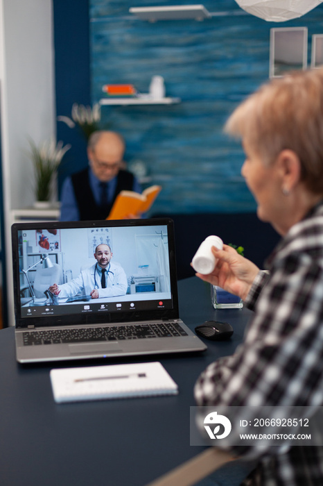 Senior pensioner listening doctor talking about treatment during online consultation. Elderly woman discussing with healthcare practitioner in the course of remote call and husband is reading a book