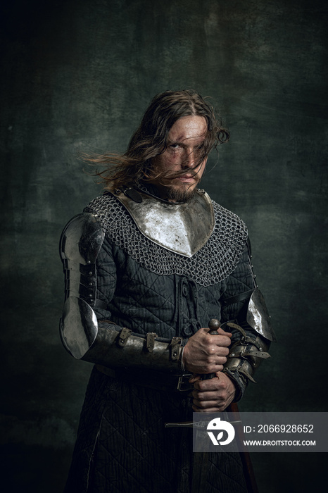 Half-length portrait of brutal seriuos man in image of medieval warrior or knight with dirty wounded face holding big sword isolated over dark background.