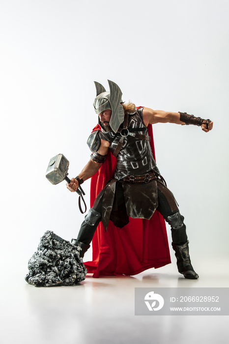 Long hair and muscular male model in leather viking’s costume with the big hammer cosplaying Thor isolated on white studio background. Full-lenght portrait. Fantasy warrior, antique battle concept.