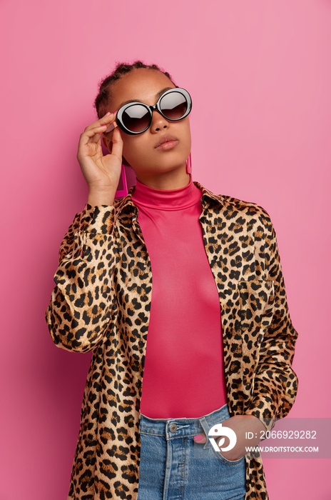 Photo of attractive fashionable woman wears sunglasses, shirt with leopard print, boasts of new purchases, being confident in herself, poses against pink background. Female in trendy clothes