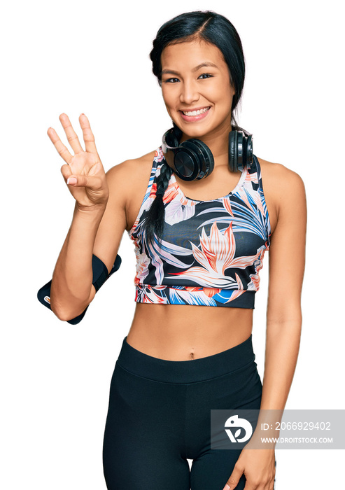 Beautiful hispanic woman wearing sportswear and headphones showing and pointing up with fingers number three while smiling confident and happy.