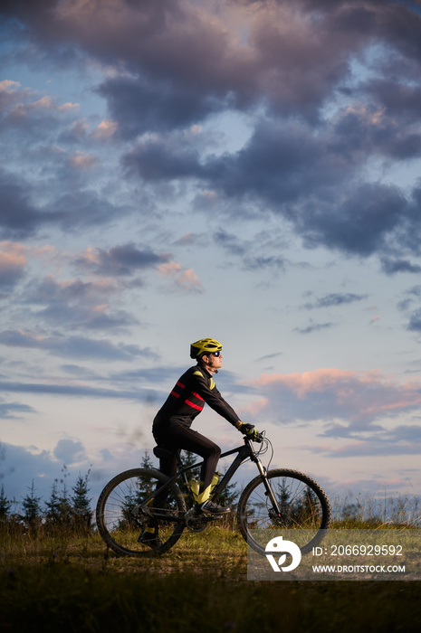 Vertical snapshot of man cyclist riding bike on sunset in the mountains. Bicyclist wearing helmet, sports glasses and uniform. Side view. Evening sky on background. Concept of active lifestyle