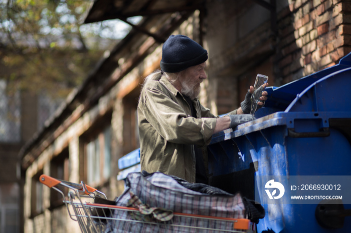 Old man collecting empty bottles to earn money. Homeless man putting empty bottles in bag in shopping cart.