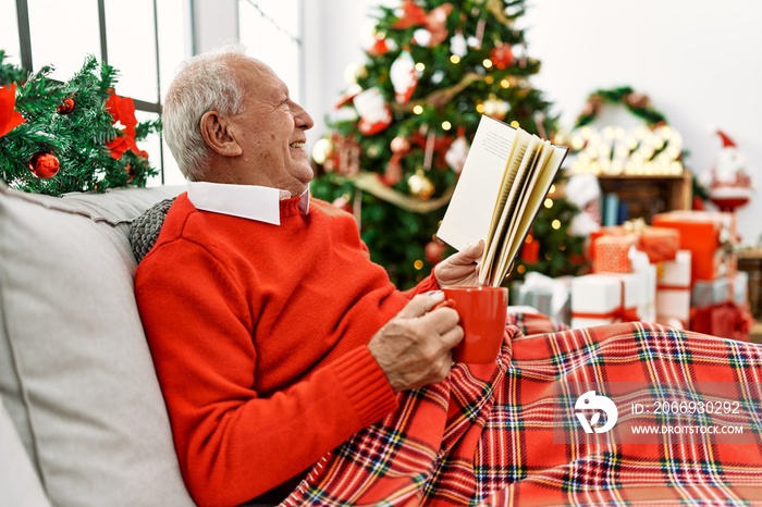 Senior grey-haired man drinking coffee and reading book celebrating christmas at home.