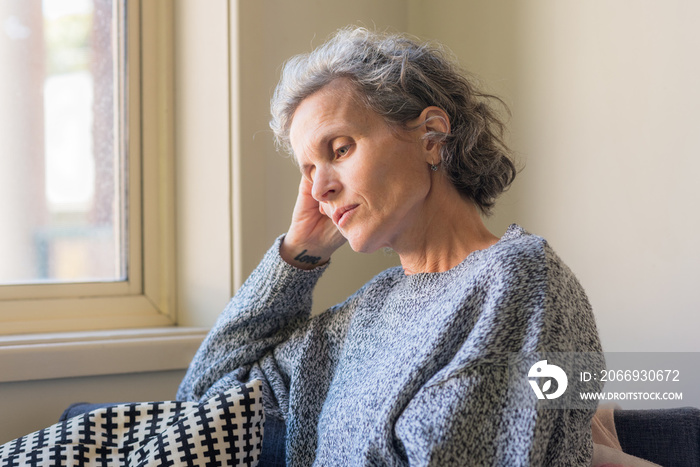 Closeup of middle aged woman with grey hair resting head on hand next to window (selective focus)