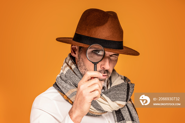 Male detective with magnifying glass, wearing fashionable scarf and brown hat, posing on yellow studio background.