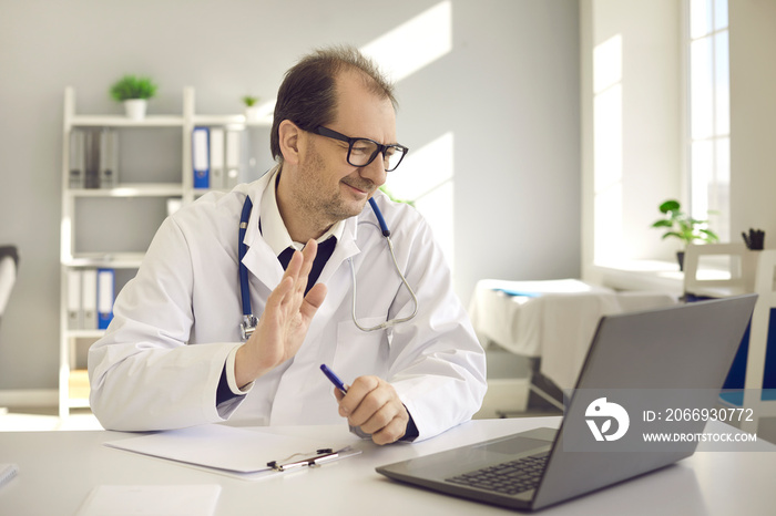 Happy physician sitting at office desk and waving hello at laptop computer greeting patient in video conference meeting. Online doctor, telemedicine consultation, virtual visit to hospital concept