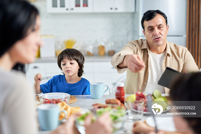 Middle aged father chatting with children, using tablet pc while eating indoors. Latin family enjoying meal together, sitting at the table in kitchen at home