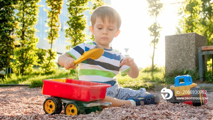 Portrait of adorable 3 years old toddler boy playing with toy truck with trailer on the playground at park. Child digging and building from sand