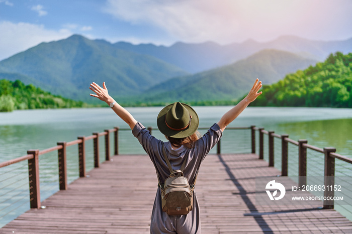 Traveler girl backpacker with open raised arms standing alone on pier and staring at lake and mountains. Enjoying free moment life and serene quiet peaceful atmosphere in nature. Back view