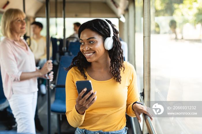 Cheerful black lady using smartphone wearing headphones in bus