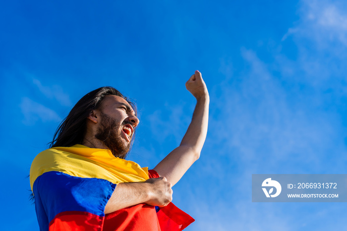 Latino Colombian man at a Colombian protest dressed in a Colombian flag, shouting with his fist raised in the air.