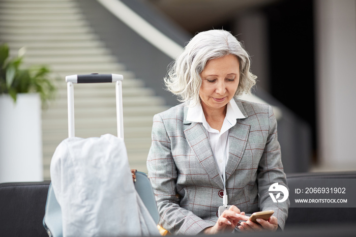 Smiling gray-haired Asian lady in checkered jacket sitting in airport and checking messenger on phone