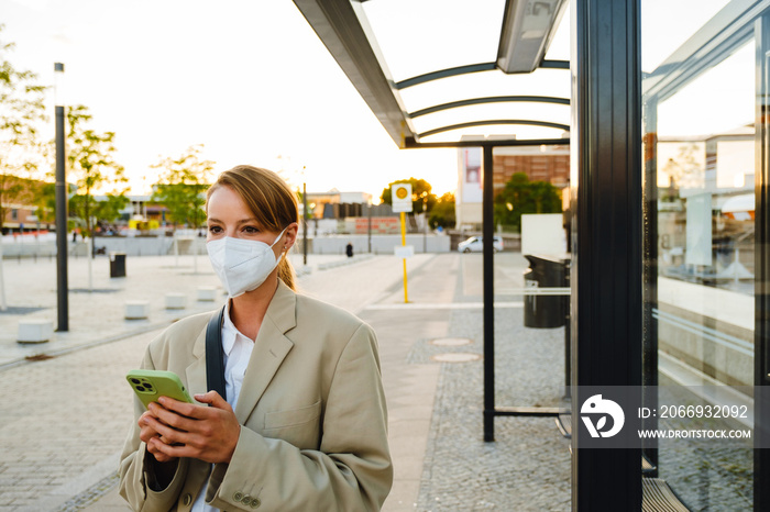 Young woman in facial mask using cellphone standing at bus station