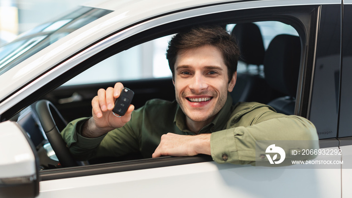 Happy young Caucasian man showing key from auto, sitting inside car salon, buying brand new automobile at dealership