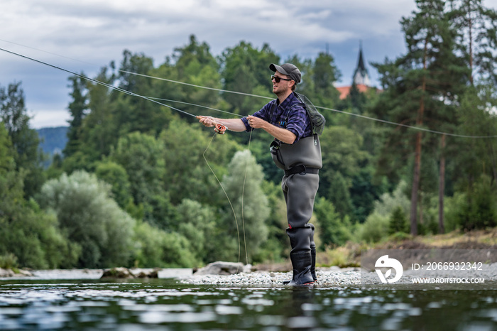 Angler mit Wathose und Fliegenrute im Wasser beim Angeln bei Sonne im klaren Fluss stehend und werfend