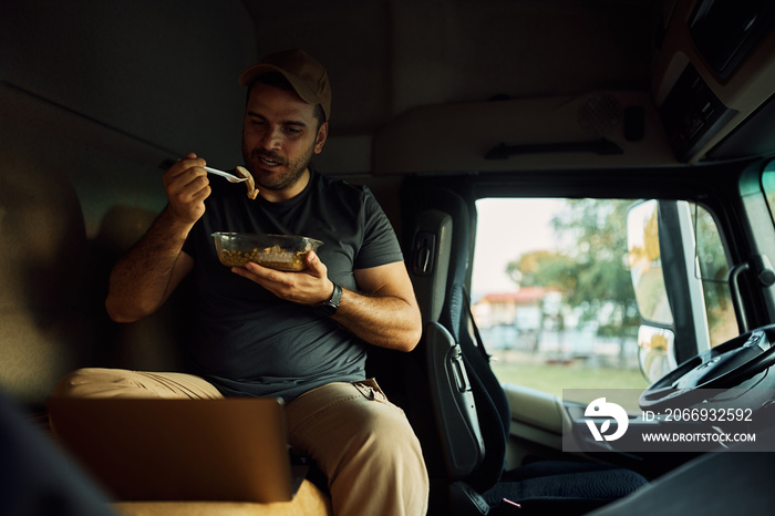 Truck driver eating lunch while using laptop in cabin.