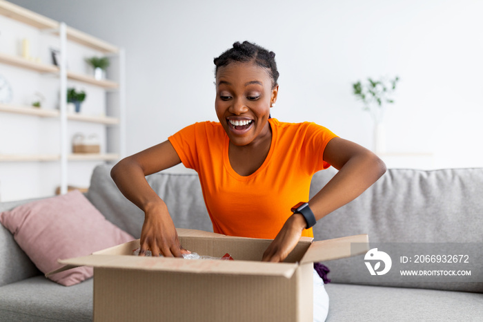 Emotional black female customer receiving long-awaited package, opening carton box at home, satisfied with purchase