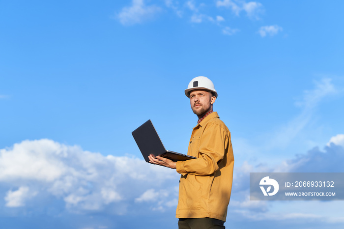 Beautiful caucasian bearded building contractor, foreman or engineer in white helmet and orange jacket using laptop at construction site at sunny day with blue cloudy sky. High quality image