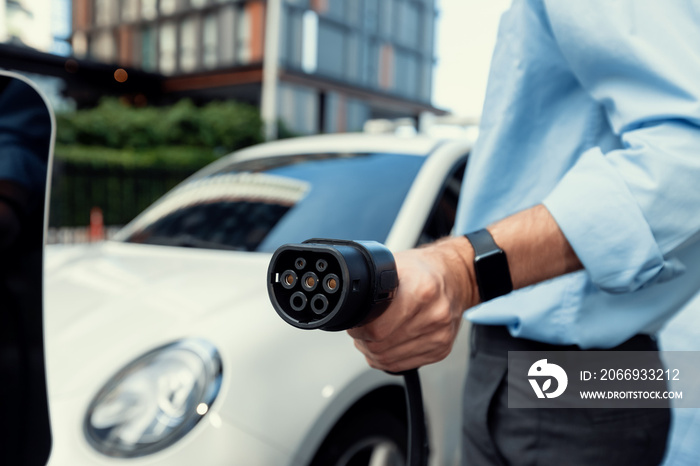 Closeup progressive man holding EV charger plug from public charging station for electric vehicle with background of residential building as concept eco-friendly sustainability energy car concept.