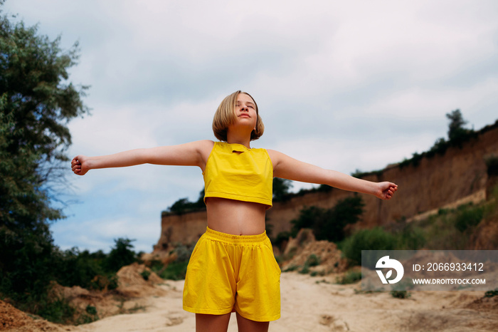 A young girl with her eyes closed, in a yellow summer suit, shorts and a tank top, stands among the sandy hills and spreads her arms to the sides.