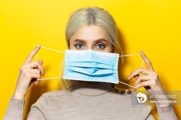 Studio portrait of young blonde girl holding a medical flu mask on face, on yellow background.