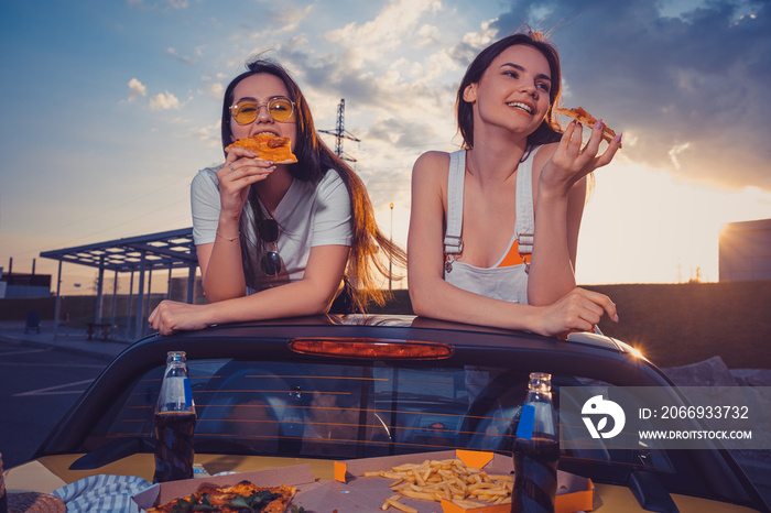 Charming women are eating pizza and smiling while posing in yellow car with french fries and soda in glass bottles on trunk. Fast food. Copy space