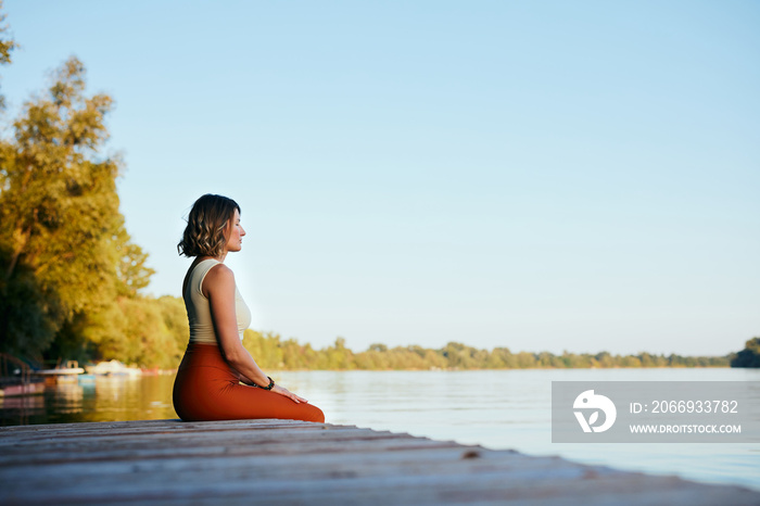 A middle-aged woman is relaxing on the dock and doing yoga breathing exercises. She put her feet in the water.