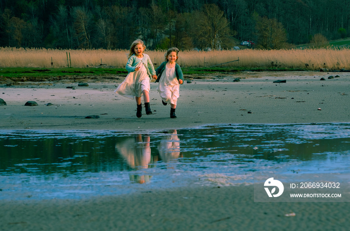 Two charming girls in dresses run along the seashore, holding hands and laughing with happiness