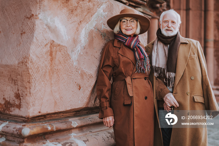 Portrait of senior bearded man and his wife standing near old building. They looking at camera and smiling