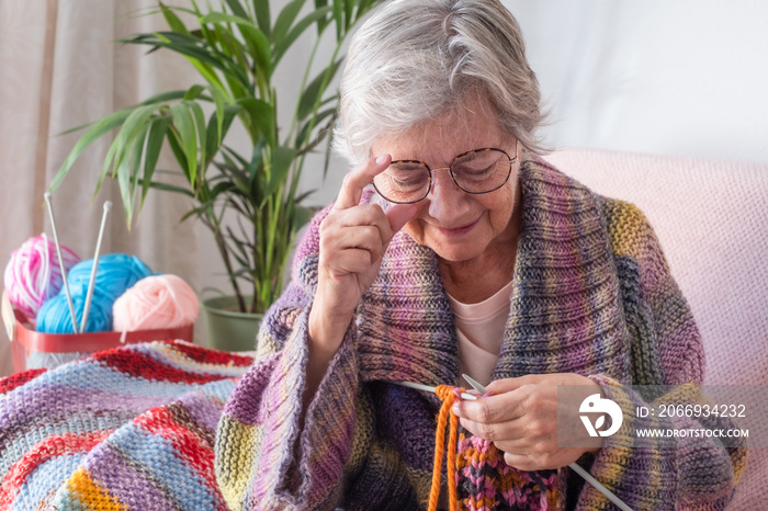 Senior mature woman sitting on sofa at home while knitting and looking at her work in progress. Hobby, retirement, relax concept for elderly female grandmother wearing glasses