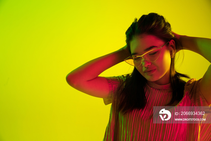 Beautiful female half-length portrait isolated on yellow studio background in neon light. Young emotional woman. Human emotions, facial expression concept. Dance in eyeglasses and wireless headphones.