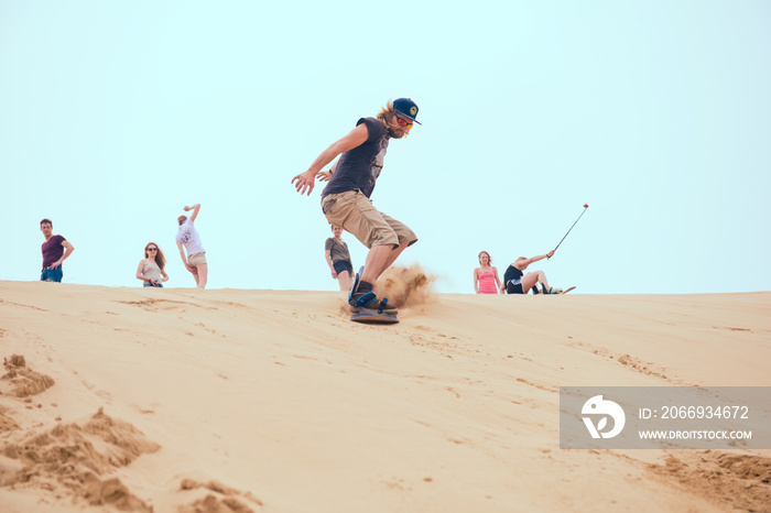 A man in beige shorts and black t-shirt sand boarding down the sand dune with few other people watching