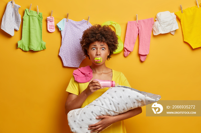 Caring loving mother keeps nipple in mouth, feeds baby with milk bottle, holds newborn on hands, busy with nursing and domestic chores, stands against yellow background, has surprised expression