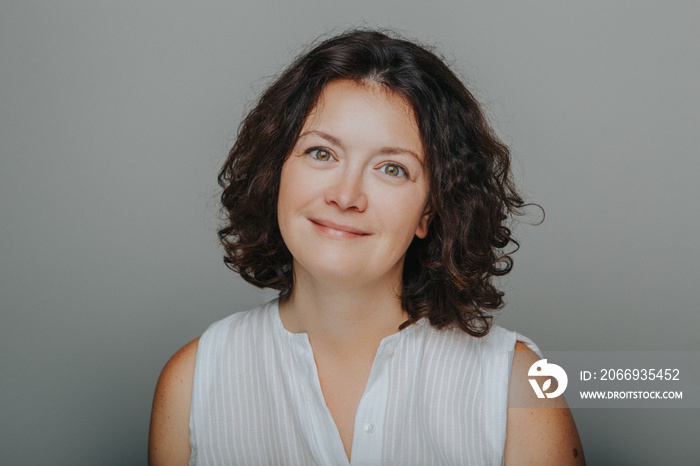 Studio head shot portrait of beautiful smiling brunette Caucasian middle aged woman with curly dark hair. Casual style model real person in white shirt.