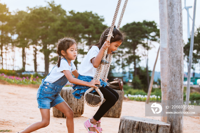 Happy asian child girl having fun to play on wooden swings with her sister in playground with beautiful nature